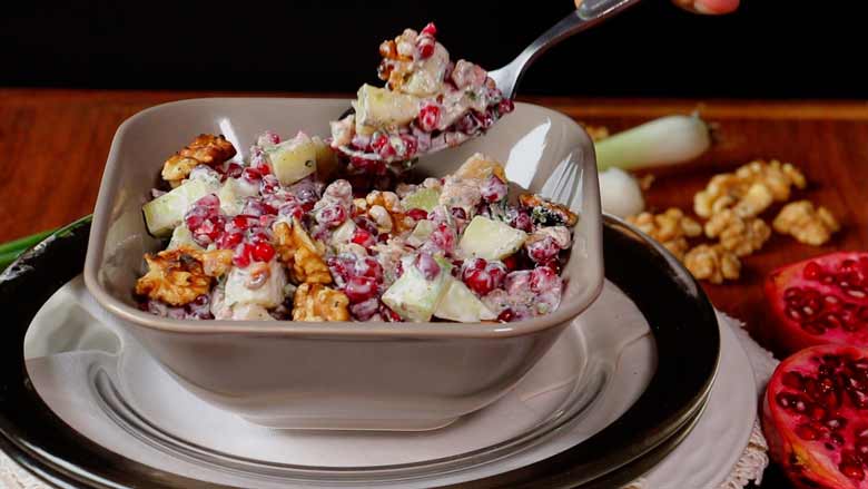 A side shot of Konkani Cucumber and Pomegranate Salad garnished with toasted California walnuts in a square grey ceramic bowl with pomegranates, California walnuts, and spring onions in the background - Easy Konkani Cucumber and Pomegranate Salad Recipe
